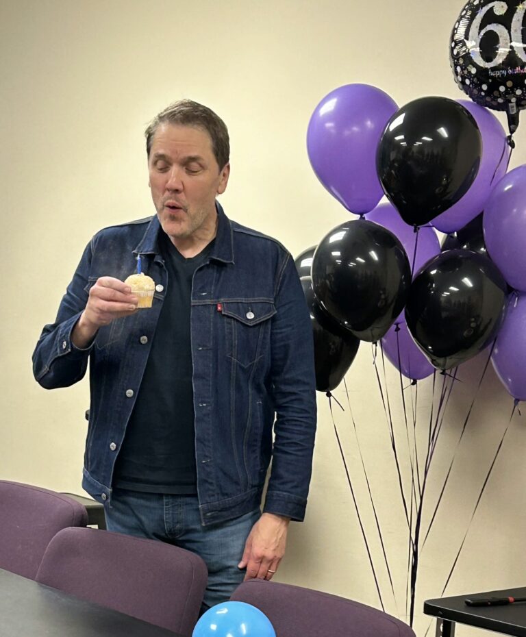 Photo of Brett blowing out a candle on a cupcake with balloons behind him